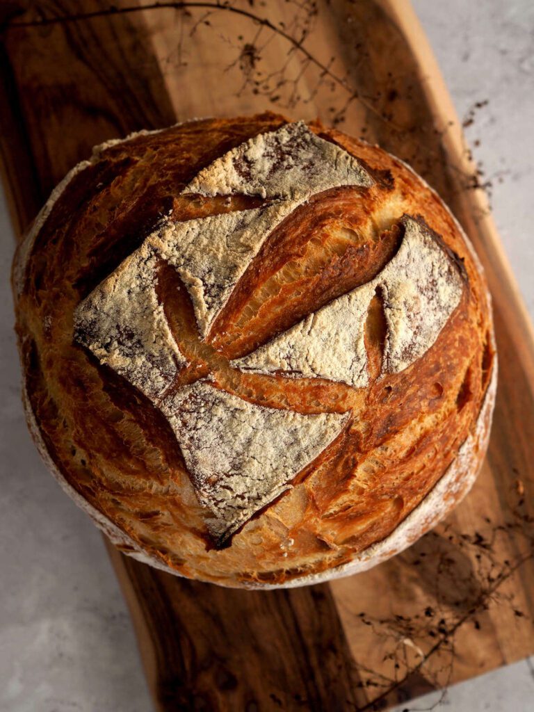 A rustic loaf of ancient bread with a rough, imperfect crust, dusted with flour, resting on a dark, weathered surface. The bread has deep cracks and an uneven shape, evoking an earthy, handmade feel.