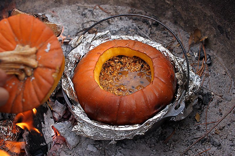 A rustic whole pumpkin with a golden-brown, slightly charred exterior, stuffed with a savory mixture of grains and nuts, cooking over an open fire in a bed of ash and foil, with a hand lifting the pumpkin’s lid nearby.