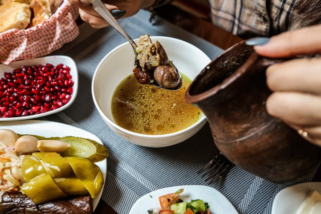 A bowl of traditional Piti soup with rich golden broth, chunks of lamb, chickpeas, and dried fruits being lifted with a spoon, surrounded by side dishes including pickles, pomegranate seeds, and fresh bread.