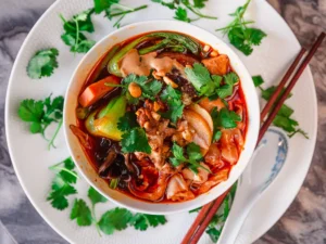 Bowl of Sichuan fiery pork and lotus stew with a deep red chili-infused broth, topped with tender pork, lotus root slices, vegetables, and fresh cilantro, served with chopsticks and a porcelain spoon on a white plate.