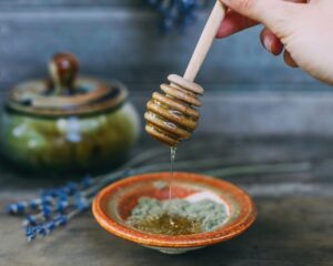 Rustic honey scene with a wooden honey dipper drizzling golden honey into a ceramic bowl, surrounded by dried lavender and an antique clay pot, set against a dark, moody background.