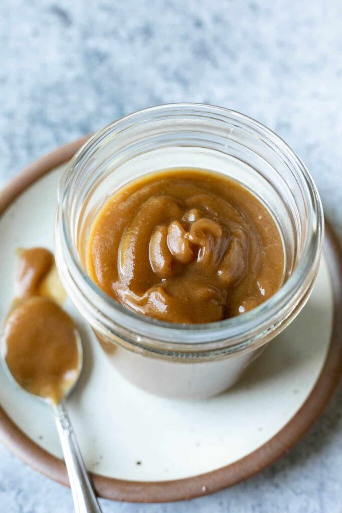 A glass jar filled with thick, golden-brown date relish with a glossy texture, sitting on a ceramic plate with a spoonful of relish beside it.