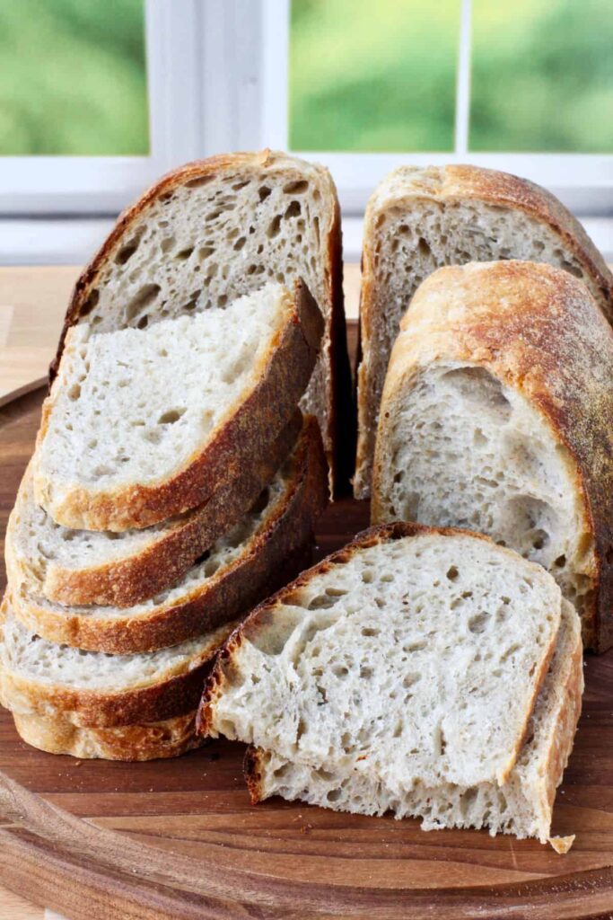 A freshly baked golden-brown loaf of bread resting on a modern stone countertop, draped with soft linen, with sunlight streaming in through a window. A glass jar of honey sits nearby, surrounded by scattered saffron flowers and spring blooms, evoking a rustic atmosphere.