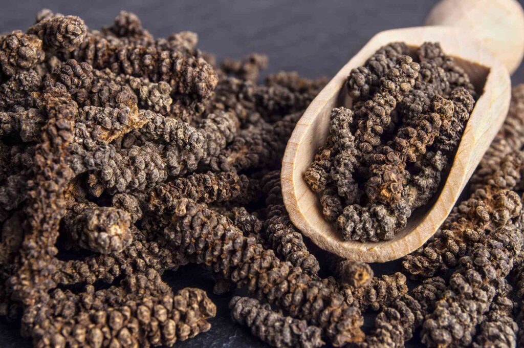 Rustic wooden bowl filled with dried long pepper (Piper longum), with some pieces scattered around on a weathered wooden surface.