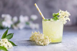 A refreshing glass of pale yellow floral elixir with ice, garnished with elderflowers and fresh mint, accompanied by a yellow-striped straw, set against a soft, textured grey background with blurred white blossoms.