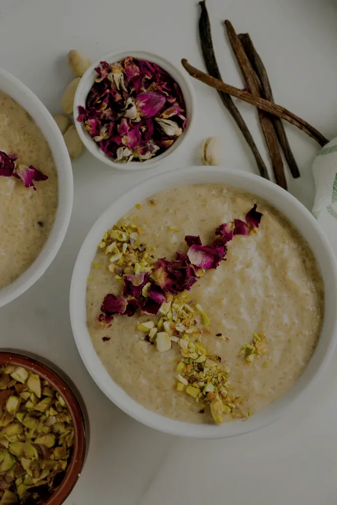 A creamy rose-infused rice pudding garnished with crushed pistachios and vibrant edible flowers, served in a white bowl on a rustic surface with vanilla pods and dried roses nearby.