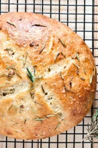 A golden-brown rustic loaf of salt-crusted herbed bread, sprinkled with fresh rosemary and coarse sea salt, cooling on a wire rack.