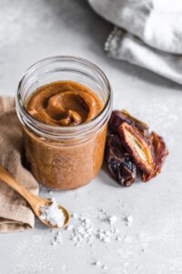 A glass jar of savoury Silphium & Date relish with a rich brown color, placed on a light textured surface. Whole and halved dates are arranged beside the jar, and a wooden spoon with coarse salt rests nearby, with scattered salt crystals adding a rustic touch.