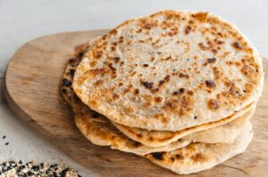 Stack of golden-brown seeded flatbreads with a crisp, bubbly surface, resting on a wooden board with scattered sesame and nigella seeds nearby.