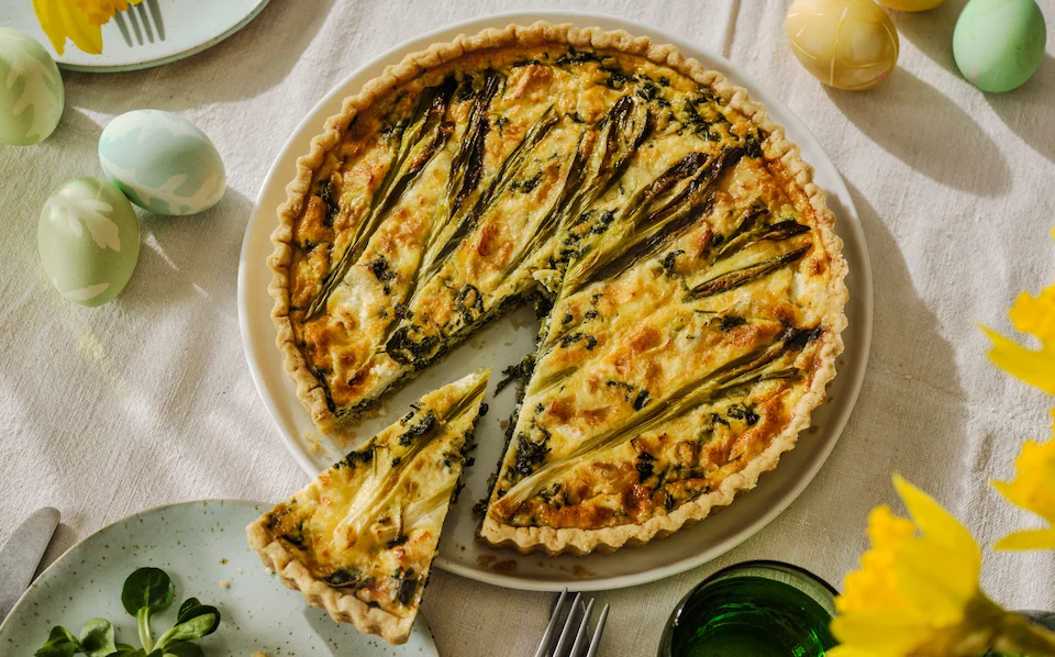 Golden spring cheese and herb tart with wild garlic and nettles, served on a white plate with a slice cut out, surrounded by pastel-colored eggs and yellow flowers on a linen tablecloth.