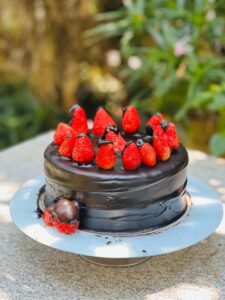 A rich, double-layer chocolate fudge cake coated in dark chocolate ganache, topped with vibrant red strawberries and a pair dipped in chocolate, set outdoors on a stone slab with soft natural light.